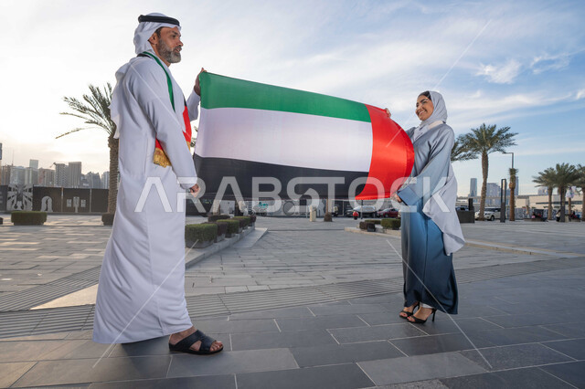 ذكرى استقلال دولة الإمارات العربية المتحدة، اليوم الوطني الإماراتي 2 ديسمبر، رجل عربي خليجي اماراتي يرتدي الكندورة والغترة يحتفل بالعيد الوطني برفقة زوجته في النهار، التجول في الشوارع بايماءات السعادة والفخر والاعتزاز، حب الوطن والانتماء اليه، رفع علم وراية الوطن