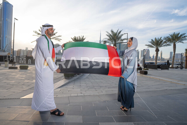 UAE Independence Day, UAE National Day, December 2, an Emirati Gulf Arab man wearing a kandura and ghutra celebrating the National Day with his wife during the day, walking in the streets with gestures of happiness, pride and honor, love of the homeland and belonging to it, raising the flag and banner of the homeland