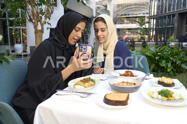 Gestures of happiness and pleasure in gathering friends, sharing food and sweets, developing social relationships, chatting and having a good time with friends, two Arab Gulf Emirati women sitting at a table in a cafe, two Saudi friends exchanging conversation.