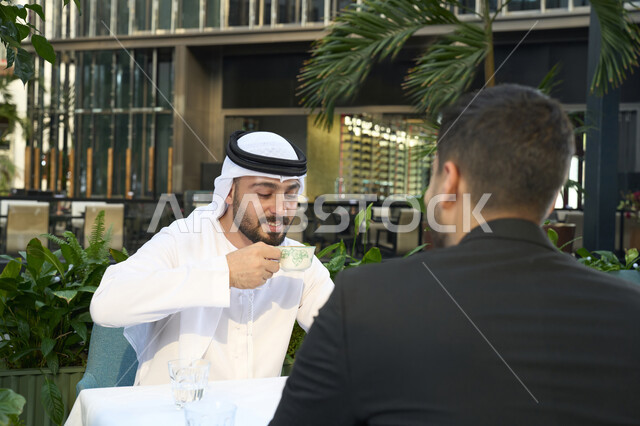Sitting and enjoying hot drinks and warm winter herbs, expressions of pleasure and happiness, an Arab Gulf Emirati businessman in a business meeting with a new client wearing a formal suit, having a good time with friends, a close-up of an Emirati young man wearing a white kandura and ghutra sitting in a cafe.