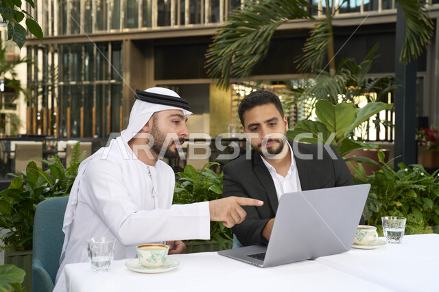 دمج العمل مع التقنية والتكنولوجيا ،مشاركة المشروبات الساخنة والقهوة، تبادل الاحاديث بين الأصدقاء، رجل عربي خليجي اماراتي يرتدي الكندورة والغترة البيضاء يجلس مع صديقه بالبدلة الرسمية بايماءات السعادة والسرور ، استخدام الحاسوب المحمول والعمل عن بعد، زملاء عمل عرب .