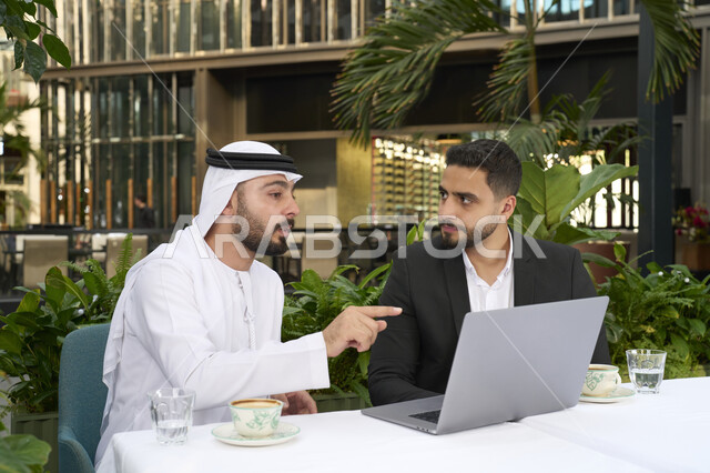 استخدام الحاسوب المحمول والعمل عن بعد، مشاركة المشروبات الساخنة والقهوة، تبادل الاحاديث بين الأصدقاء، رجل عربي خليجي اماراتي يرتدي الكندورة والغترة البيضاء يجلس مع صديقه بالبدلة الرسمية بايماءات السعادة والسرور ، زملاء عمل عرب .