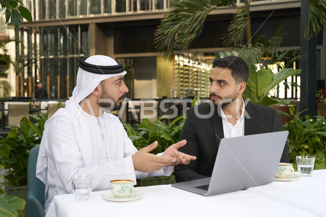 مناقشة مهام العمل في المقهى، إدارة شؤون الأعمال وتنظيمها عبر الأجهزة التقنية الحديثة، مشاركة المشروبات الساخنة والقهوة، تبادل الاحاديث بين الأصدقاء، رجل عربي خليجي اماراتي يرتدي الكندورة والغترة البيضاء يجلس مع صديقه بالبدلة الرسمية بايماءات السعادة والسرور .