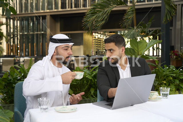 زملاء عمل عرب , قضاء وقت ممتع في الخارج، إدارة شؤون الأعمال وتنظيمها عبر الأجهزة التقنية الحديثة، مشاركة المشروبات الساخنة والقهوة، تبادل الاحاديث بين الأصدقاء،رجل عربي خليجي اماراتي يرتدي الكندورة والغترة البيضاء يجلس مع صديقه بالبدلة الرسمية في المقهى بايماءات السعادة والسرور .