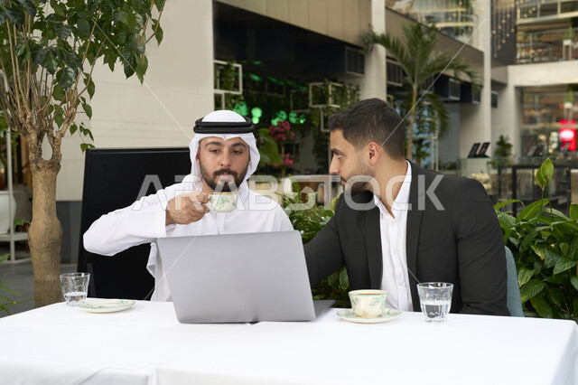 مشاركة المشروبات الساخنة والقهوة، تبادل الاحاديث بين الأصدقاء، قضاء وقت ممتع في الخارج، التسلية والترفيه في العطلة، استخدام الحاسوب المحمول ، انجاز المهام عن بعد ، رجل عربي خليجي اماراتي يرتدي الكندورة والغترة البيضاء يجلس مع صديقه بالبدلة الرسمية في المقهى بايماءات السعادة والسرور .