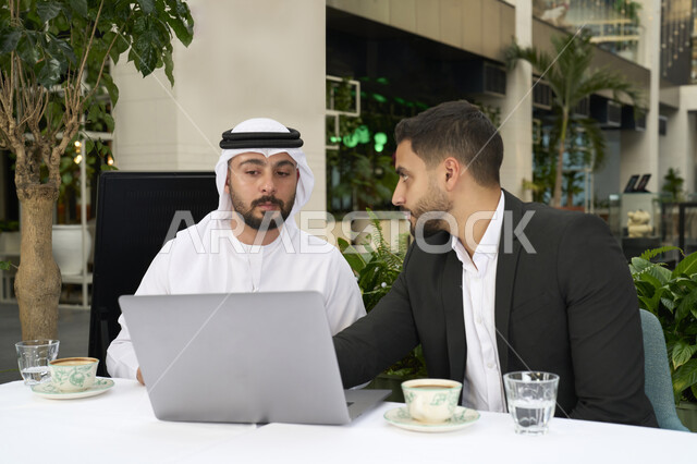 حوارات ونقاشات عمل , إدارة شؤون الأعمال وتنظيمها عبر الأجهزة التقنية الحديثة، مشاركة المشروبات الساخنة والقهوة، قضاء وقت ممتع في الخارج، تبادل الاحاديث بين الأصدقاء،رجل عربي خليجي اماراتي يرتدي الكندورة والغترة البيضاء يجلس مع صديقه بالبدلة الرسمية في المقهى بايماءات السعادة والسرور .