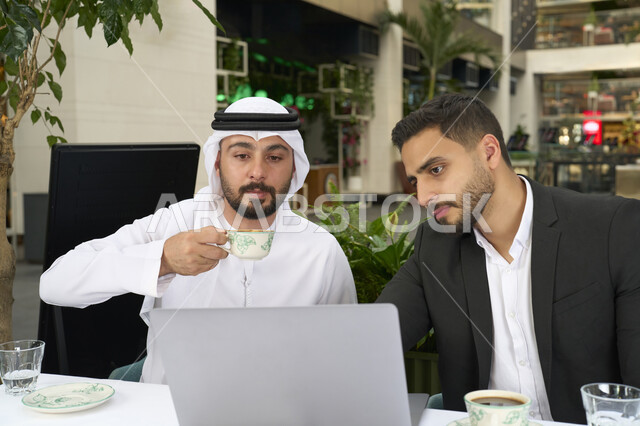 انجاز المهام عن بعد ، مشاركة المشروبات الساخنة والقهوة، تبادل الاحاديث بين الأصدقاء، قضاء وقت ممتع في الخارج، التسلية والترفيه في العطلة، استخدام الحاسوب المحمول ، رجل عربي خليجي اماراتي يرتدي الكندورة والغترة البيضاء يجلس مع صديقه بالبدلة الرسمية في المقهى بايماءات السعادة والسرور .
