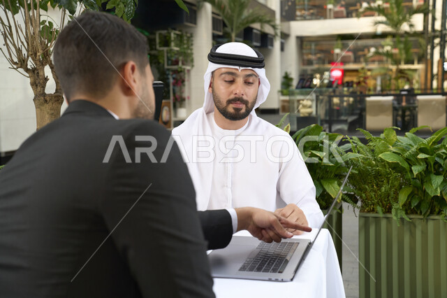 اجتماع عمل لتطوير المشاريع و الاستثمارات ،عمل جماعي مشترك ،رجل اعمال عربي خليجي اماراتي في اجتماع عمل مع عميل جديد يرتدي البدلة الرسمية، استخدم جهاز الحاسوب المحمول ، صورة مقربة لشاب إماراتي يرتدي الكندورة والغترة البيضاء ، انجاز المشاريع والمهام في الكافيه.