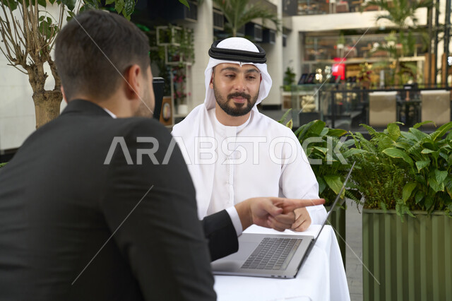 An Emirati work team and a man in a formal suit in a business meeting, using blank paper to record work tasks, an Emirati Gulf Arab businessman in a business meeting with a new client, using a laptop, a close-up of an Emirati young man wearing a white kandora and ghutra, completing projects and tasks in a café.