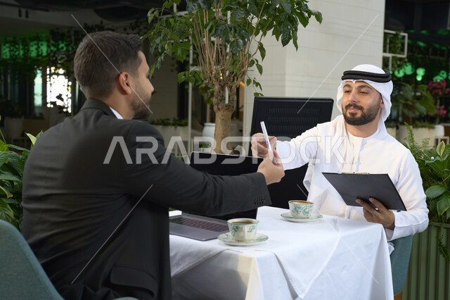 مهن ووظائف اماراتية ، استخدام الاوراق البيضاء فى  تدوين مهام العمل ،عمل جماعي مشترك ،رجل اعمال عربي خليجي اماراتي في اجتماع عمل مع عميل جديد يرتدي البدلة الرسمية، استخدم جهاز الحاسوب المحمول ، صورة مقربة لشاب إماراتي يرتدي الكندورة والغترة البيضاء ، انجاز المشاريع والمهام في الكافيه.