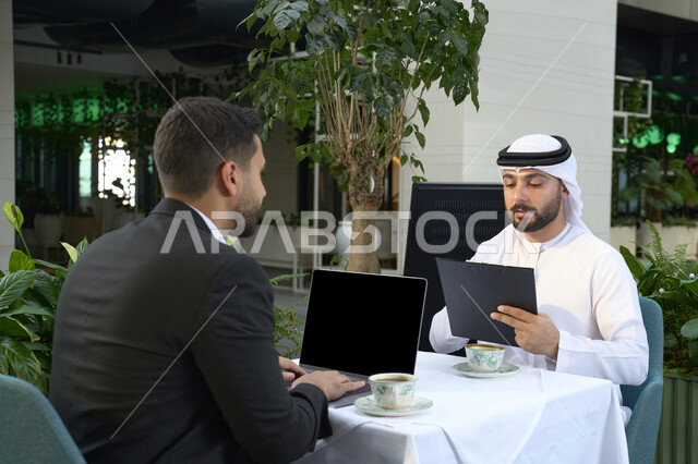 ايماءات الجدية والتركيز فى العمل ، رجل اعمال عربي خليجي اماراتي في اجتماع عمل مع عميل جديد يرتدي البدلة الرسمية، استخدم جهاز الحاسوب المحمول ، صورة مقربة لشاب إماراتي يرتدي الكندورة والغترة البيضاء ، انجاز المشاريع والمهام في الكافيه.