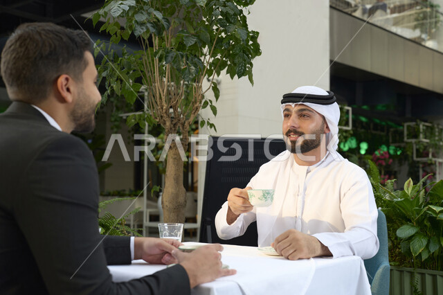 تبادل الاحاديث بين الأصدقاء، مشاركة المشروبات الساخنة والقهوة،  قضاء وقت ممتع في الخارج، رجل عربي خليجي اماراتي يرتدي الكندورة والغترة البيضاء يجلس مع صديقه بالبدلة الرسمية في المقهى بايماءات السعادة والسرور .