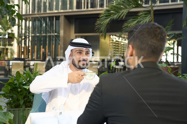 ايماءات السعادة والسرور، مشاركة المشروبات الساخنة والقهوة، قضاء وقت ممتع في الخارج، تبادل الاحاديث بين الأصدقاء،رجل عربي خليجي اماراتي يرتدي الكندورة والغترة البيضاء يجلس مع صديقه بالبدلة الرسمية في المقهى  .