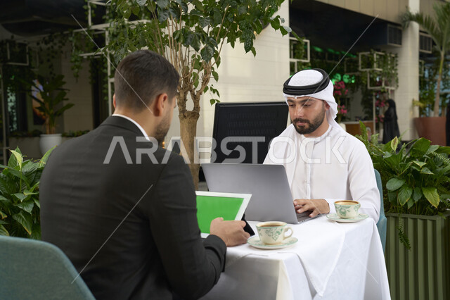 استخدام التقنيات الحديثة فى العمل ، انجاز المشاريع والمهام في الكافيه ، تدوين مهام العمل ، رجل اعمال عربي خليجي اماراتي في اجتماع عمل مع عميل جديد يرتدي البدلة الرسمية، استخدم جهاز الحاسوب المحمول ، صورة مقربة لشاب إماراتي يرتدي الكندورة والغترة البيضاء يجلس فى المقهى .