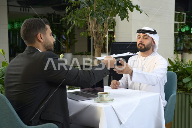 Completing projects and tasks in a cafe, writing down work tasks, an Arab Gulf Emirati businessman in a business meeting with a new client wearing a formal suit, using a laptop, a close-up of a young Emirati man wearing a white kandura and ghutra sitting in a cafe.