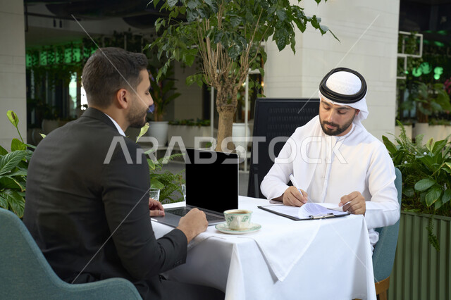 إدارة شؤون الأعمال وتنظيمها عبر الأجهزة التقنية الحديثة ، استخدم جهاز الحاسوب المحمول لانجاز المشاريع والمهام في الكافيه ، قضاء وقت ممتع مع الأصدقاء, صورة مقربة لشاب عربي خليجي إماراتي يرتدي الكندورة والغترة البيضاء يجلس مع رجل اعمال بالبدلة الرسمية في المقهى .