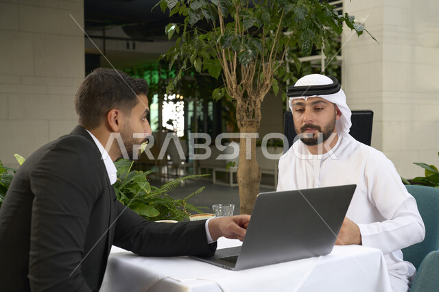 مفهوم التكنولوجيا وإستخدام أجهزة تقنية حديثة ، استخدم جهاز الحاسوب المحمول لانجاز المشاريع والمهام في الكافيه ، قضاء وقت ممتع مع الأصدقاء, صورة مقربة لشاب عربي خليجي إماراتي يرتدي الكندورة والغترة البيضاء يجلس مع رجل اعمال بالبدلة الرسمية في المقهى .