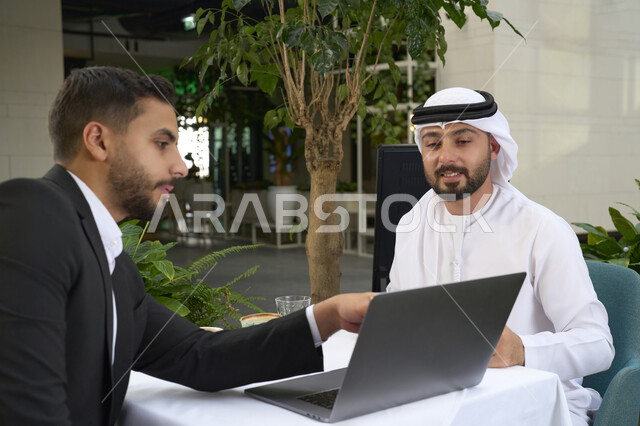 استخدم جهاز الحاسوب المحمول لانجاز المشاريع والمهام في الكافيه، ايماءات الجدية والتركيز فى العمل ، قضاء وقت ممتع مع الأصدقاء, صورة مقربة لشاب عربي خليجي إماراتي يرتدي الكندورة والغترة البيضاء يجلس مع رجل اعمال بالبدلة الرسمية في المقهى .