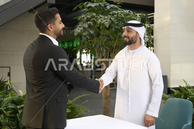 Handshaking and making business agreements, an Emirati Gulf Arab businessman with an Arab businessman wearing a formal suit, sitting in a cafe, a meeting of business colleagues, partnership and cooperation, following up on business and achieving goals.