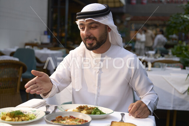 تعابير السرور والسعادة ، تناول الغداء في المقهى , قضاء أوقات ممتعة في المقهى، رجل عربي خليجي اماراتي يرتدي الكندورة والغترة البيضاء يجلس على مائدة الطعام و يشير اليه ، الاستمتاع بالأنشطة الترفيهية, مطاعم و كافيهات في الإمارات العربية المتحدة .