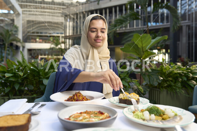 مطاعم و كافيهات في الإمارات ،  تناول الغداء في المطعم, الاستمتاع بالأنشطة الترفيهية, امرأة عربية خليجية إماراتية محجبة ترتدي الزي الكاجوال تجلس على المائدة و تنظر إلى الكاميرا بإيماءات السعادة و السرور, قضاء الأوقات الممتعة في الخارج .
