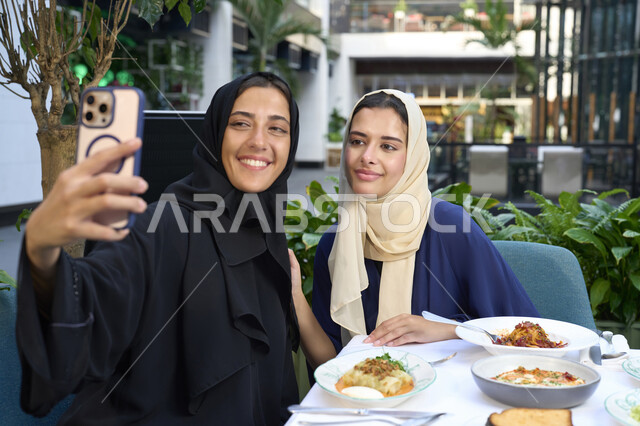 استخدام التقنيات الحديثة ،تعابير السعادة والسرور بلمة الرفاق، التسامر والدردشة وقضاء وقت ممتع مع الأصدقاء، كوفي شوب راقي بالامارات، امرأتين عربيتين خليجيتين اماراتيتين تجلسان على طاولة في المقهى، صديقتين سعوديتين تتبادلان الحديث .
