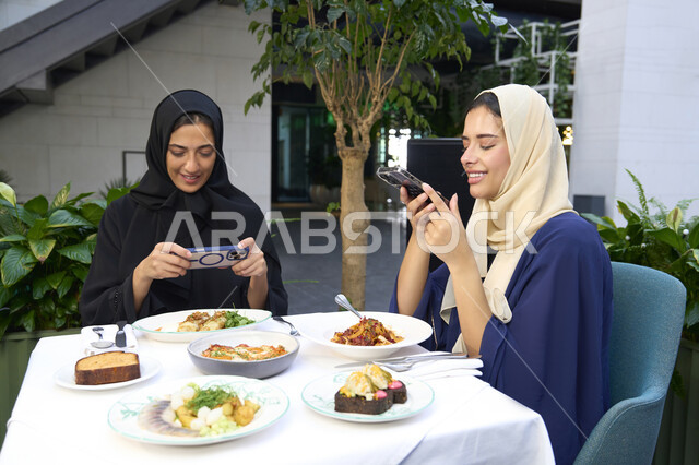 توثيق اللحظات التذكارية ، تطوير العلاقات الإجتماعية، التسامر والدردشة وقضاء وقت ممتع مع الأصدقاء، كوفي شوب راقي بالامارات، تعابير السعادة والسرور بلمة الرفاق، امرأتين عربيتين خليجيتين اماراتيتين تجلسان على طاولة في المقهى، صديقتين سعوديتين تتبادلان الحديث .