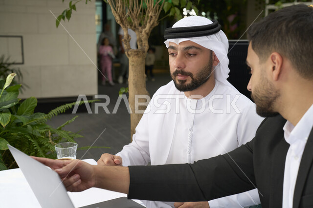 Gestures of seriousness and focus at work, using the laptop to complete projects and tasks in the café, spending quality time with friends, a close-up of a young Emirati Gulf Arab man wearing a white kandura and ghutra sitting with a businessman in a formal suit in the café.