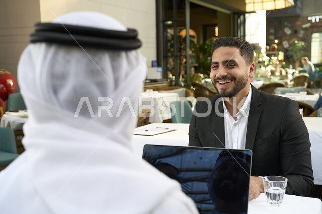 تعابير السرور والسعادة ، استخدم جهاز الحاسوب المحمول لانجاز المشاريع والمهام في الكافيه، قضاء وقت ممتع مع الأصدقاء, صورة مقربة لشاب عربي خليجي إماراتي يرتدي الكندورة والغترة البيضاء يجلس مع رجل اعمال بالبدلة الرسمية في المقهى .