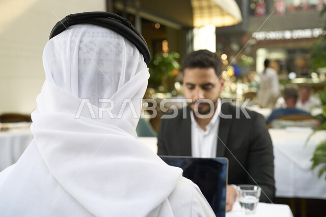 اجتماع عمل فى المقهى ،التكنولوجيا والتقننيات الحديثة ، استخدم جهاز الحاسوب المحمول لانجاز المشاريع والمهام في الكافيه، قضاء وقت ممتع مع الأصدقاء, صورة مقربة لشاب عربي خليجي إماراتي يرتدي الكندورة والغترة البيضاء يجلس مع رجل اعمال بالبدلة الرسمية .