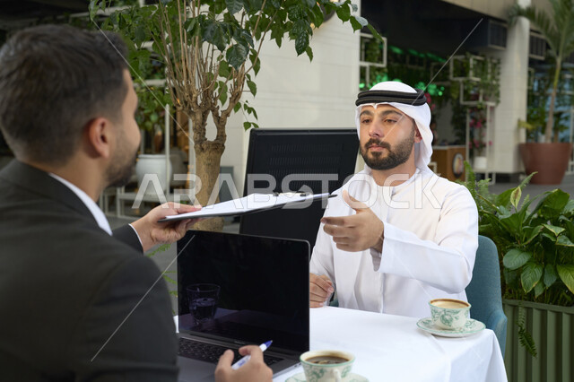 اجتماع عمل فى المقهى، استخدم جهاز الحاسوب المحمول لانجاز المشاريع والمهام في الكافيه،قضاء وقت ممتع مع الأصدقاء, صورة مقربة لشاب عربي خليجي إماراتي يرتدي الكندورة والغترة البيضاء يجلس مع صديقة بالبدلة الرسمية .