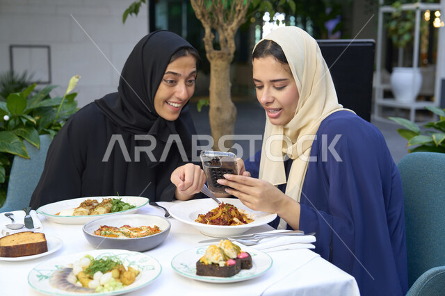 مشاركة الطعام والحلويات ،  تعابير السعادة والسرور بلمة الرفاق، تطوير العلاقات الإجتماعية، التسامر والدردشة وقضاء وقت ممتع مع الأصدقاء، امرأتين عربيتين خليجيتين اماراتيتين تجلسان على طاولة في المقهى، صديقتين سعوديتين تتبادلان الحديث .
