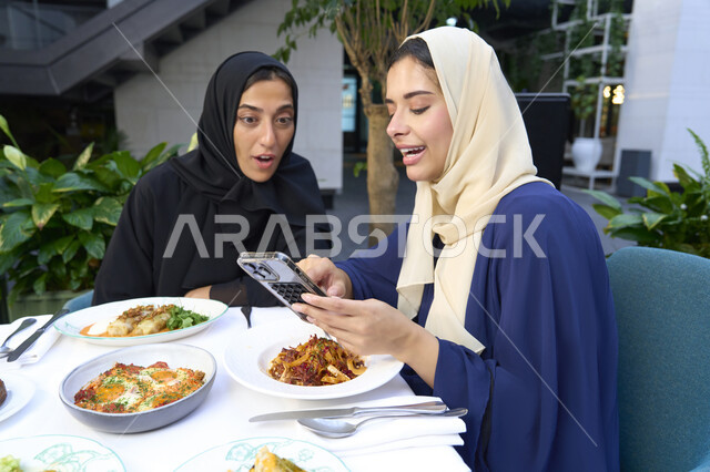 النظر الى شئ ما فى الجوال ،  تعابير السعادة والسرور بلمة الرفاق، تطوير العلاقات الإجتماعية، التسامر والدردشة وقضاء وقت ممتع مع الأصدقاء، امرأتين عربيتين خليجيتين اماراتيتين تجلسان على طاولة في المقهى، صديقتين سعوديتين تتبادلان الحديث .