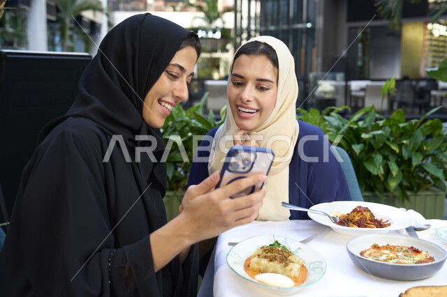 Developing social relationships, chatting and having a good time with friends, a high-end coffee shop in the Emirates, expressions of happiness and pleasure at a gathering of friends, two Emirati Gulf Arab women sitting at a table in a cafe, two Saudi friends exchanging conversation.