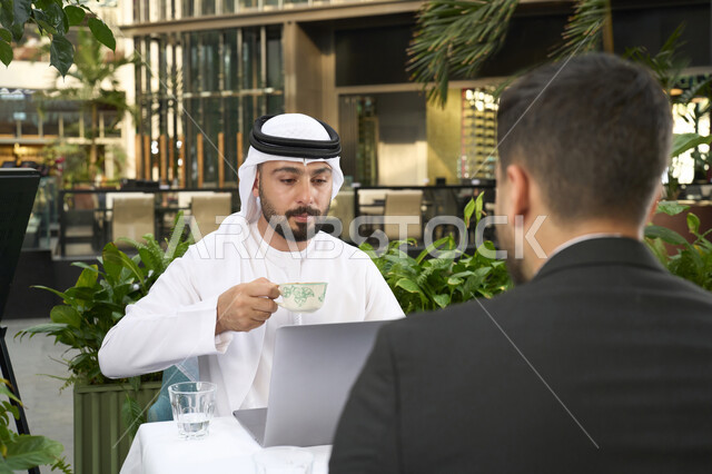 Hosting businessmen in the UAE, using a laptop to complete projects and tasks in the cafe, spending quality time with friends, a close-up of a young Emirati Gulf Arab man wearing a white kandura and ghutra sitting with his friend in a formal suit in the cafe.