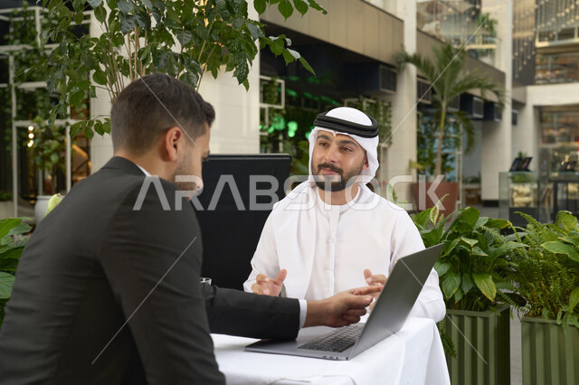 الحوار والنقاش بين زملاء العمل، استخدم جهاز الحاسوب المحمول لانجاز المشاريع والمهام في الكافيه،قضاء وقت ممتع مع الأصدقاء, صورة مقربة لشاب عربي خليجي إماراتي يرتدي الكندورة والغترة البيضاء يجلس مع صديقة بالبدلة الرسمية في المقهى .