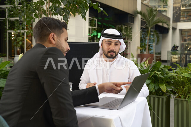 استخدم جهاز الحاسوب المحمول لانجاز المشاريع والمهام في الكافيه، الحوار والنقاش بين زملاء العمل، قضاء وقت ممتع مع الأصدقاء, صورة مقربة لشاب عربي خليجي إماراتي يرتدي الكندورة والغترة البيضاء يجلس مع صديقه في المقهى .