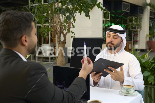 الحوار والنقاش بين زملاء العمل، استخدم جهاز الحاسوب المحمول لانجاز المشاريع والمهام في الكافيه، قضاء وقت ممتع مع الأصدقاء, صورة مقربة لشاب عربي خليجي إماراتي يرتدي الكندورة والغترة البيضاء يجلس مع صديقه في المقهى .
