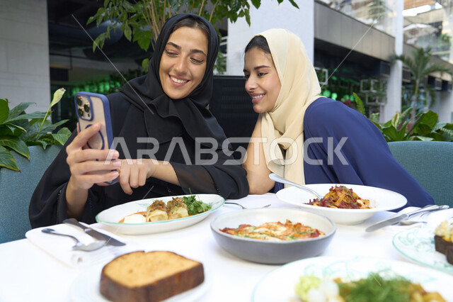 Chatting, having a good time with friends, taking souvenir photos, a classy coffee shop in the Emirates, developing social relationships, expressions of happiness and pleasure at a gathering of friends, two Emirati Gulf Arab women sitting at a table in a cafe, two Saudi friends exchanging talk.
