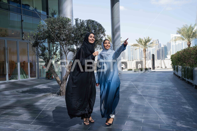 الاشارة والتوجية الى مكان معين ، الوقوف بثقة في شوارع المدينة، الاستمتاع والتجول واستنشاق الهواء النقي في وقت النهار، صديقتان عربيتان خليجيتان سعوديتان ترتديان العباءات والحجاب ، زميلتان اماراتيان تنظران الى شئ ما بتعابير السرور، مفهوم الأناقة والاهتمام بالمظهر الخارجي .