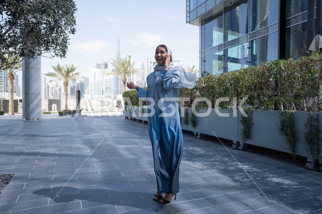 Making a phone call on a mobile phone, chatting and communicating with family, hearing news and happy surprises, taking a walk in the open air, an Emirati Gulf Arab woman having a good time in a park in the Emirates, a Saudi woman taking a walk in nature, a full-length body image.