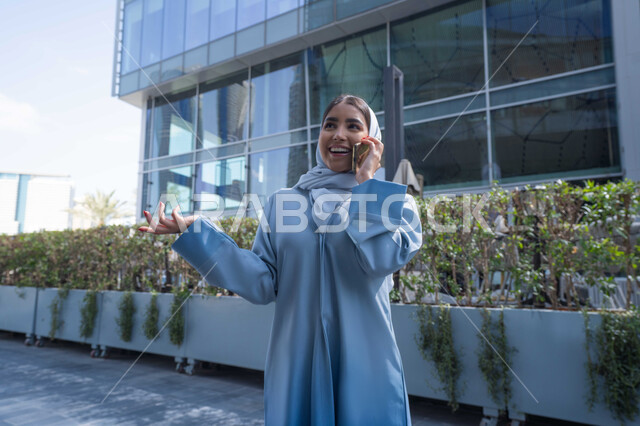 Merging life with modern technology, an Emirati woman using a mobile phone, chatting on social media, gestures of happiness when hearing good news, checking on friends, exchanging conversations with family, a smiling young Saudi Arabian Gulf woman holding a mobile phone in her hand.