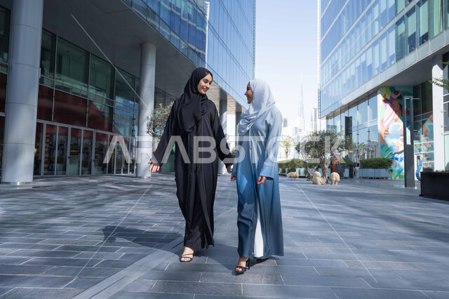 المشي فى الهواء الطلق ، الفخر والاعتزاز بالتقدم المعماري ، سيدتان اماراتيان تنظران النمو والتطور العمراني ، التجول واستنشاق الهواء النقي في الخارج, صديقتان عربيتان خليجيتان سعوديتان ترتديان العباءات والحجاب تنظران الى بعضهما بتعابير السرور في وقت النهار.