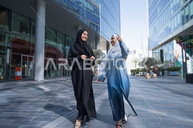 الاستمتاع بالتنزة فى شوارع المدينة ، الفخر والاعتزاز بالتقدم المعماري ، سيدتان اماراتيان تنظران النمو والتطور العمراني ، التجول واستنشاق الهواء النقي في الخارج, صديقتان عربيتان خليجيتان سعوديتان ترتديان العباءات والحجاب تنظران الى بعضهما بتعابير السرور في وقت النهار.