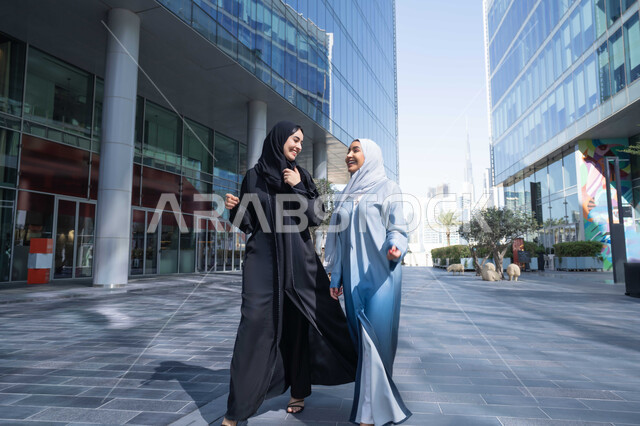 تبادل الاحاديث والاخبار بين الاصدقاء ، الفخر والاعتزاز بالتقدم المعماري ، سيدتان اماراتيان تنظران النمو والتطور العمراني ، التجول واستنشاق الهواء النقي في الخارج, صديقتان عربيتان خليجيتان سعوديتان ترتديان العباءات والحجاب تنظران الى شئ ما بتعابير السرور في وقت النهار.