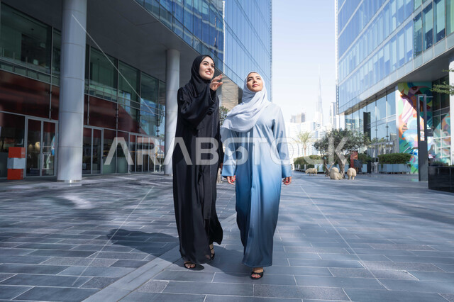 Pointing to a specific location, taking pride in architectural progress, two Emirati women looking at urban growth and development, walking around and breathing fresh air outside, two Saudi Arabian Gulf friends wearing abayas and hijab looking at something with happy expressions during the day.