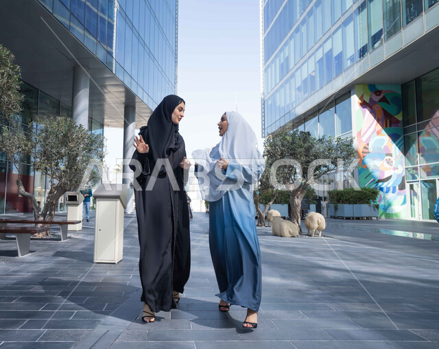 التجول واستنشاق الهواء النقي في الخارج, الفخر والاعتزاز بالتقدم المعماري ، امرأتان اماراتيان تنظر النمو والتطور العمراني ، صديقتان عربيتان خليجيتان سعوديتان ترتديان العباءات والحجاب تنظران الى بعضهما بتعابير السرور في وقت النهار.