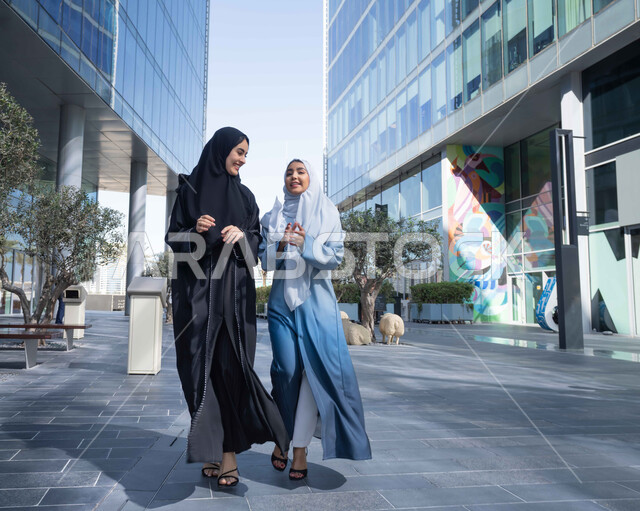 النزهة والترفيه فى العطلة الاسبوعية  ، الفخر والاعتزاز بالتقدم المعماري ، سيدتان اماراتيان تنظران النمو والتطور العمراني ، التجول واستنشاق الهواء النقي في الخارج, صديقتان عربيتان خليجيتان سعوديتان ترتديان العباءات والحجاب تنظران الى شئ ما بتعابير السرور في وقت النهار.