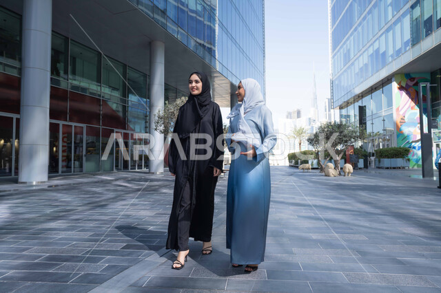 جولة مع الاصدقاء فى شوارع المدينة ، الفخر والاعتزاز بالتقدم المعماري ، سيدتان اماراتيان تنظران النمو والتطور العمراني ، التجول واستنشاق الهواء النقي في الخارج, صديقتان عربيتان خليجيتان سعوديتان ترتديان العباءات والحجاب تنظران الى شئ ما بتعابير السرور في وقت النهار.