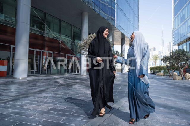 جولة مع الاصدقاء فى شوارع المدينة ، الفخر والاعتزاز بالتقدم المعماري ، سيدتان اماراتيان تنظران النمو والتطور العمراني ، التجول واستنشاق الهواء النقي في الخارج, صديقتان عربيتان خليجيتان سعوديتان ترتديان العباءات والحجاب تنظران الى شئ ما بتعابير السرور في وقت النهار.